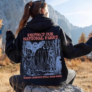 Op de afbeelding: Een zwarte hoodie met een grafische print op de achterkant met de tekst "PROTECT OUR NATIONAL PARKS" en "WHERE LEGENDS STILL ROAM FREE" in het rood, samen met blauwe illustraties van een vleermuis, een hert, een sasquatch en spookachtige figuren.