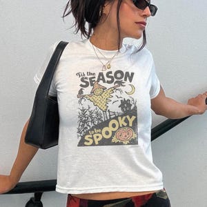 May include: A white short-sleeve t-shirt featuring a graphic print with the text 'Tis the SEASON to be SPOOKY'. The illustration depicts a scarecrow, a crescent moon, a haunted house, a crow, and a jack-o'-lantern in a field setting.