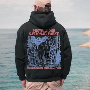 May include: A black hooded sweatshirt featuring a graphic print on the back with the text PROTECT OUR NATIONAL PARKS and WHERE LEGENDS STILL ROAM FREE. The illustration depicts a forest scene with a sasquatch, an owl, and other figures.