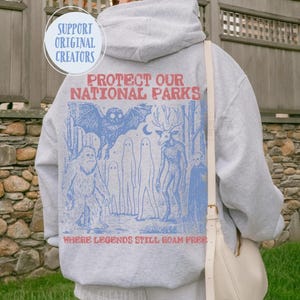 May include: A light grey hooded sweatshirt featuring a blue and red graphic on the back. The text reads "PROTECT OUR NATIONAL PARKS" and "WHERE LEGENDS STILL ROAM FREE" with illustrations of a sasquatch, an owl, ghosts, and a deer-like creature.