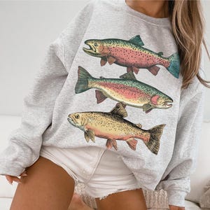 May include: A light gray sweatshirt featuring three detailed illustrations of trout. The fish are depicted in shades of pink, yellow, and green, with intricate fin and scale details. The sweatshirt is a casual, comfortable style.