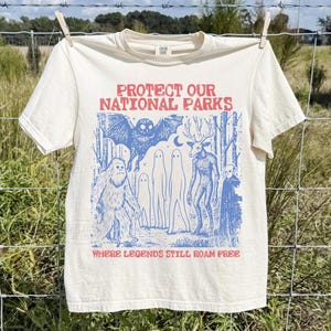 Peut inclure: Un t-shirt &agrave; manches courtes couleur cr&egrave;me avec une impression graphique bleue repr&eacute;sentant une chauve-souris, un sasquatch, des fant&ocirc;mes, une cr&eacute;ature ressemblant &agrave; un cerf et une for&ecirc;t. Le texte indique PROTECT OUR NATIONAL PARKS et WHERE LEGENDS STILL ROAM FREE en rouge.