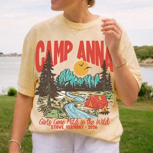Op de afbeelding: Een lichtgeel t-shirt met korte mouwen en een grafische print met de tekst CAMP ANNA, Girls Gone Mild in the Wild, en STOWE, VERMONT - 2026. Het ontwerp toont een landschap met dennenbomen, bergen, een rivier en een rode tent.