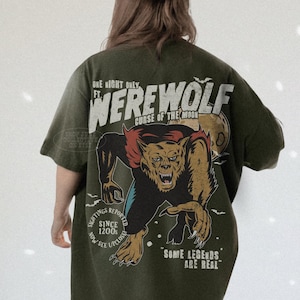 May include: An olive green t-shirt featuring a large graphic on the back of a snarling werewolf with red fur accents and a full moon. The text reads: ONE NIGHT ONLY FT. WEREWOLF CURSE OF THE MOON, SIGHTINGS REPORTED SINCE 1200S NOW SEE UPCLOSE, SOME LEGENDS ARE REAL.