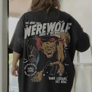 May include: A charcoal gray t-shirt featuring a large graphic on the back. The design depicts a werewolf with the text "ONE NIGHT ONLY FT. WEREWOLF CURSE OF THE MOON," "SIGHTINGS REPORTED SINCE 1200s," and "SOME LEGENDS ARE REAL" in white and red.