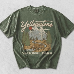 May include: A faded olive green short-sleeve t-shirt featuring a graphic print of Yellowstone National Park. The design includes a brown bison, a winding river, pine trees, mountains, and a red sun, with the text Yellowstone National Park.
