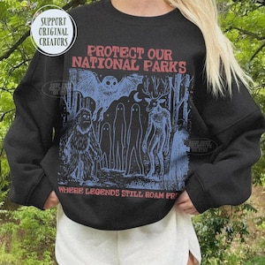 May include: A black long-sleeve sweatshirt featuring a graphic print in blue and red. The text reads PROTECT OUR NATIONAL PARKS and WHERE LEGENDS STILL ROAM FREE. The illustration depicts a forest scene with a large creature, a deer-like figure, ghosts, and a bat.