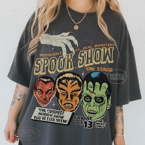 May include: A charcoal gray oversized t-shirt featuring a graphic print with the text Midnight Spook Show On Stage. The design includes a white hand illustration and three monster faces in red, orange, and green, with the text The Creepiest Horror Show You've Ever Seen and Friday Oct 13 One Night Only.