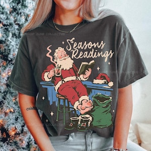May include: A charcoal gray short-sleeve t-shirt featuring a graphic of Santa Claus sitting on a stool, reading a book, and holding a steaming mug. The text "Seasons Readings" is printed above him, accompanied by a green sack and boots.