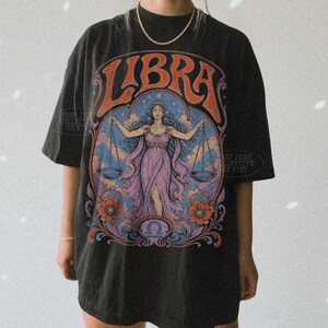 May include: A black short-sleeve t-shirt featuring a large graphic print on the front. The design displays the word LIBRA in orange letters above an illustration of a figure holding scales, surrounded by blue clouds, orange flowers, and swirls.