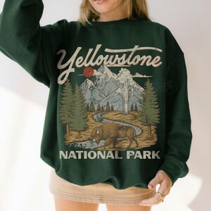 May include: A dark green crewneck sweatshirt featuring a graphic print of Yellowstone National Park. The design includes a bison, a winding river, pine trees, mountains, and a red sun, with the text Yellowstone National Park in a vintage font.