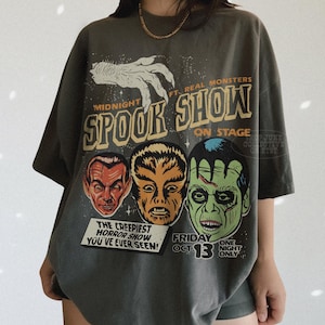 May include: A charcoal gray short-sleeve t-shirt featuring a graphic print with the text MIDNIGHT SPOOK SHOW FT. REAL MONSTERS ON STAGE. The design includes a white clawed hand, three monster faces, and the text THE CREEPIEST HORROR SHOW YOU'VE EVER SEEN!
