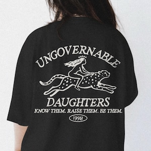 Puede incluir: Camiseta negra con un gráfico blanco en la espalda. El gráfico presenta las palabras "UNGOVERNABLE DAUGHTERS" sobre una ilustración de una mujer montando un guepardo. Debajo, las palabras "KNOW THEM. RAISE THEM. BE THEM." y el año "1998".