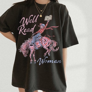 May include: Dark grey t-shirt featuring the text "Well Read Woman" in pink script. The graphic shows a person on a pink horse holding a book. The person is wearing a red shirt and blue jeans.