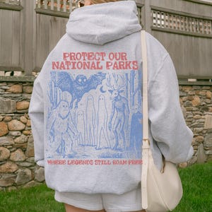 May include: A light gray hooded sweatshirt featuring a blue graphic print on the back. The text reads PROTECT OUR NATIONAL PARKS and WHERE LEGENDS STILL ROAM FREE. The illustration depicts a bat, a sasquatch, ghosts, and a deer-headed creature.
