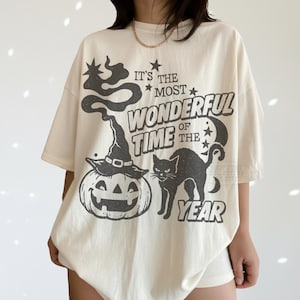 Op de afbeelding: Een off-white t-shirt met korte mouwen en een donkergrijze grafische print van een pompoen met een heksenhoed, een zwarte kat en de tekst "IT'S THE MOST WONDERFUL TIME OF THE YEAR" met sterren en een halve maan ontwerp.