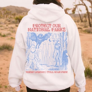 May include: A white hooded sweatshirt featuring a blue graphic print on the back. The design depicts a forest scene with a large owl, a sasquatch, two ghosts, a deer-like creature, and a figure. Text reads: PROTECT OUR NATIONAL PARKS WHERE LEGENDS STILL ROAM FREE.