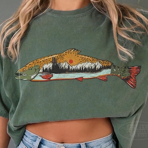 Puede incluir: Camiseta verde con una ilustraci&oacute;n de un pez que incorpora una escena de monta&ntilde;a y puesta de sol. El dise&ntilde;o del pez presenta detalles en naranja, rojo y marr&oacute;n. La camiseta se combina con vaqueros de mezclilla azul claro.