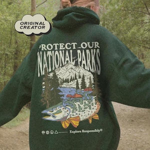 May include: A dark green hooded sweatshirt featuring a back graphic with the text PROTECT OUR NATIONAL PARKS and Explore Responsibly. The design includes a mountain landscape, pine trees, a river, and a large fish. A cloud icon says ORIGINAL CREATOR.