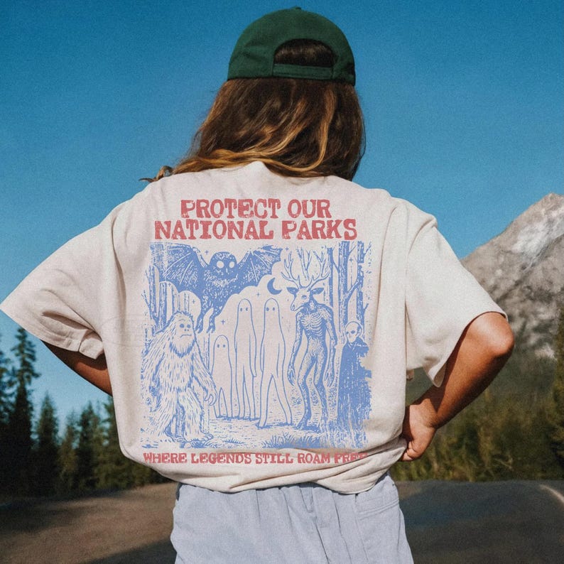 May include: A beige short-sleeve t-shirt featuring a blue graphic print on the back. The text reads "PROTECT OUR NATIONAL PARKS" and "WHERE LEGENDS STILL ROAM FREE" above and below illustrations of a bat, ghosts, a deer, and a sasquatch.