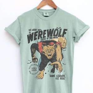 May include: A sage green short-sleeve t-shirt featuring a graphic of a werewolf with the text "ONE NIGHT ONLY FT. WEREWOLF CURSE OF THE MOON", "SIGHTINGS REPORTED SINCE 1200s NOW SEE UPCLOSE", and "SOME LEGENDS ARE REAL" with a full moon and bats.
