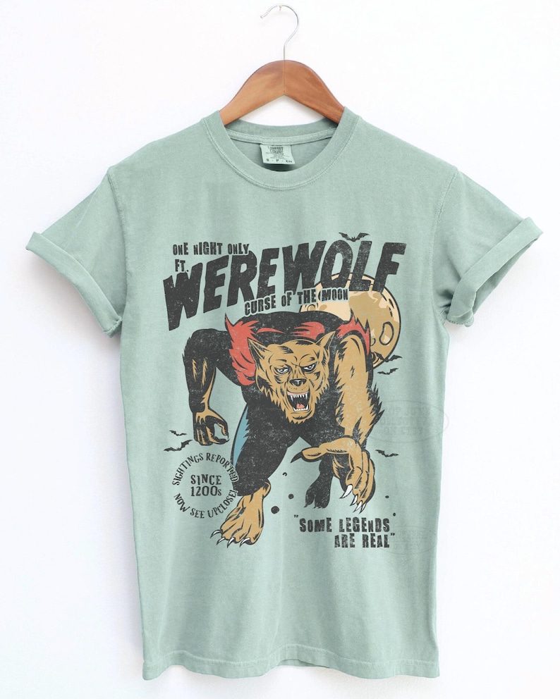 May include: A sage green short-sleeve t-shirt featuring a graphic of a werewolf with the text "ONE NIGHT ONLY FT. WEREWOLF CURSE OF THE MOON", "SIGHTINGS REPORTED SINCE 1200s NOW SEE UPCLOSE", and "SOME LEGENDS ARE REAL" with a full moon and bats.
