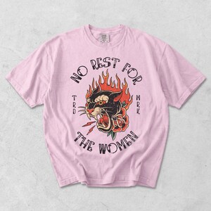 May include: Pale pink t-shirt with a vintage tattoo-style design. The design features a black panther with flames, a rose, and the words "NO REST FOR THE WOMEN".