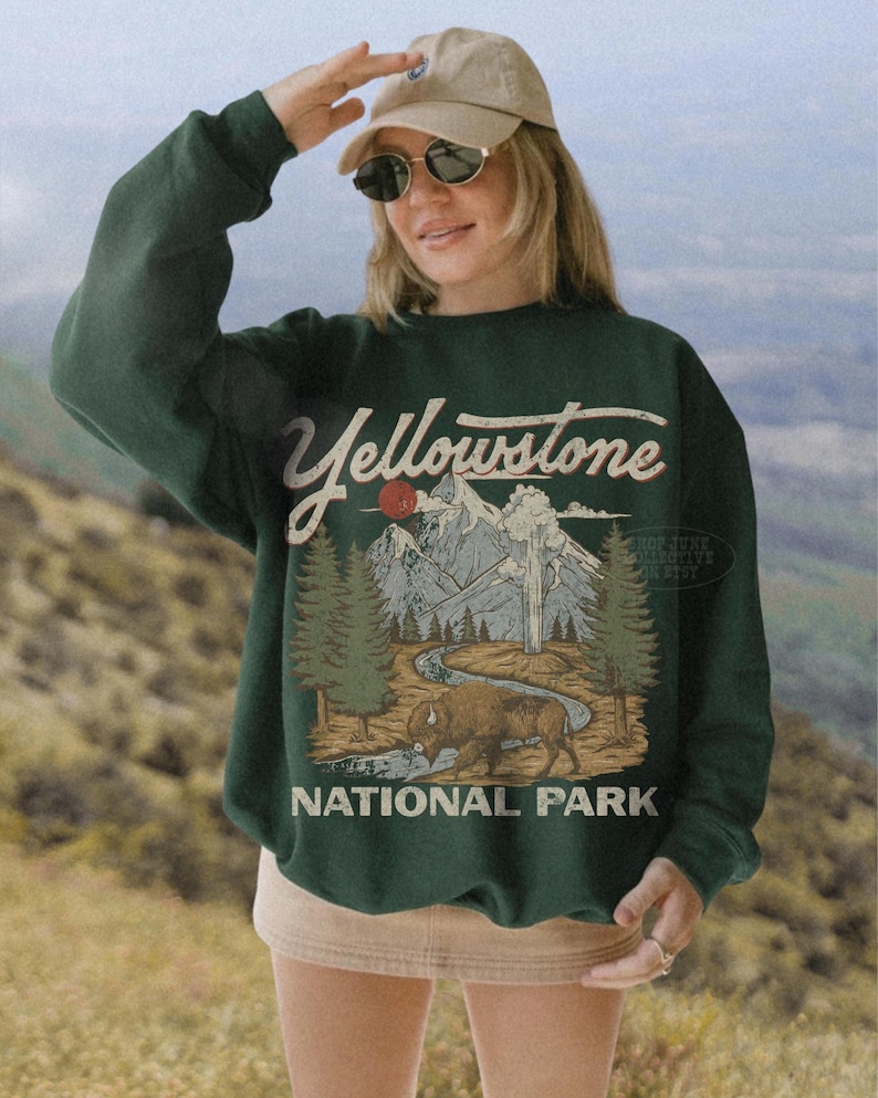May include: A dark green long-sleeve sweatshirt featuring a graphic print of a mountain landscape, pine trees, a winding river, and a bison. The text Yellowstone National Park is printed in a vintage-style font above and below the illustration.
