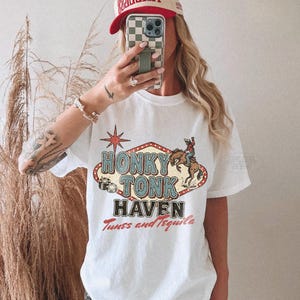 Pode incluir: Camiseta branca com um gr&aacute;fico vintage "Honky Tonk Haven" com um cowboy em um cavalo selvagem. A camisa tamb&eacute;m tem o texto "Tunes and Tequila". Um bon&eacute; de beisebol vermelho e bege com a palavra "TIMELESS" &eacute; usado.
