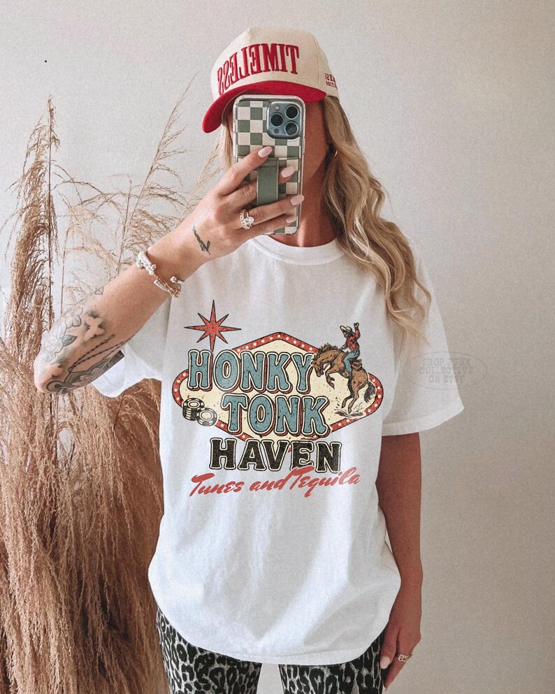 Pode incluir: Camiseta branca com um gr&aacute;fico vintage "Honky Tonk Haven" com um cowboy em um cavalo selvagem. A camisa tamb&eacute;m tem o texto "Tunes and Tequila". Um bon&eacute; de beisebol vermelho e bege com a palavra "TIMELESS" &eacute; usado.