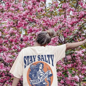 May include: A light yellow t-shirt with a graphic of a mermaid with the text "Stay Salty Siren Lounge Take No Shit" printed on the back.