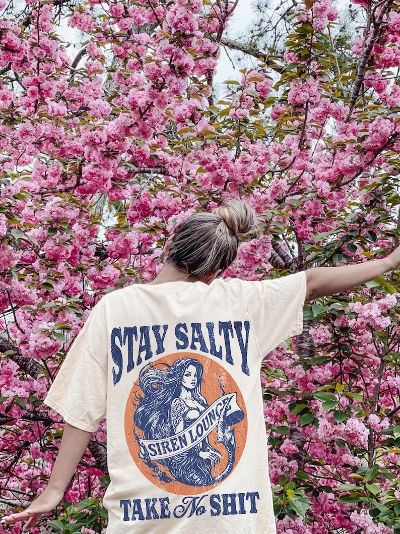 May include: A light yellow t-shirt with a graphic of a mermaid with the text "Stay Salty Siren Lounge Take No Shit" printed on the back.