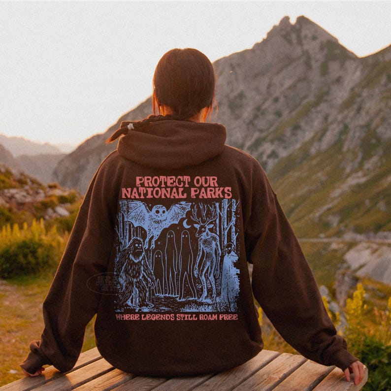 May include: A dark brown hooded sweatshirt featuring a graphic print on the back. The text reads "PROTECT OUR NATIONAL PARKS" and "WHERE LEGENDS STILL ROAM FREE" in pink, with a blue illustration of a forest, a bat, a sasquatch, and spirits.