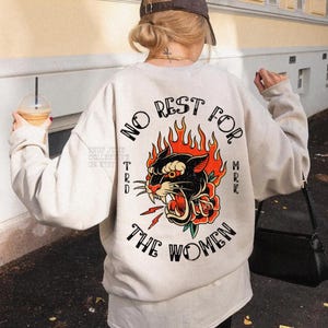 May include: Cream-colored sweatshirt with a graphic on the back featuring a black panther with red flames and a rose, and the text "NO REST FOR THE WOMEN". The sweatshirt is a casual style.