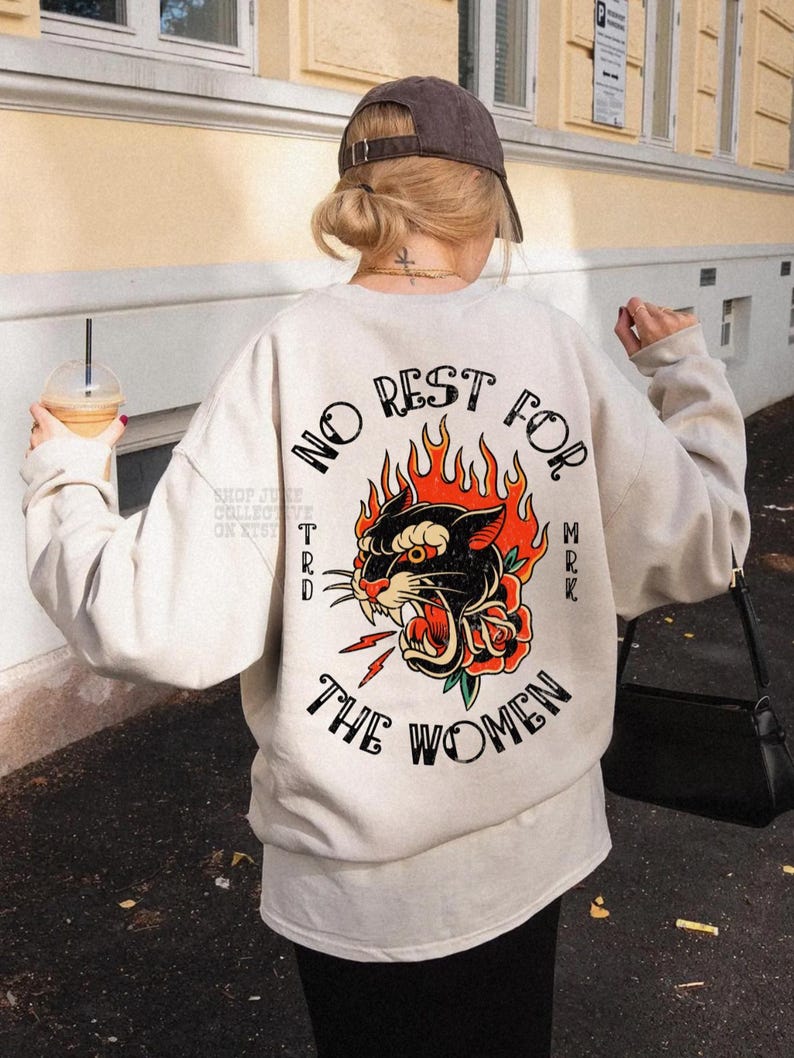 May include: Cream-colored sweatshirt with a graphic on the back featuring a black panther with red flames and a rose, and the text "NO REST FOR THE WOMEN". The sweatshirt is a casual style.