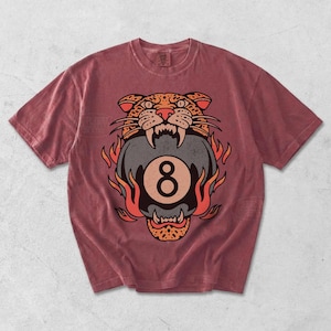 May include: A maroon t-shirt featuring a graphic of a tiger head biting an eight ball, surrounded by flames. The tiger's head is detailed with orange and black spots, and the eight ball has a large number eight. The shirt has a crew neck and short sleeves.