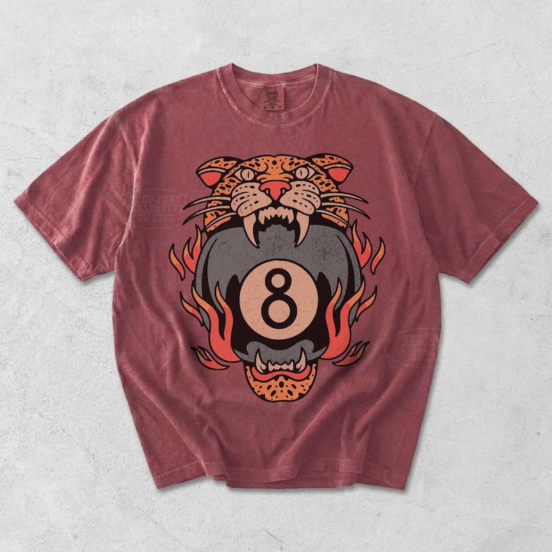 May include: A maroon t-shirt featuring a graphic of a tiger head biting an eight ball, surrounded by flames. The tiger's head is detailed with orange and black spots, and the eight ball has a large number eight. The shirt has a crew neck and short sleeves.