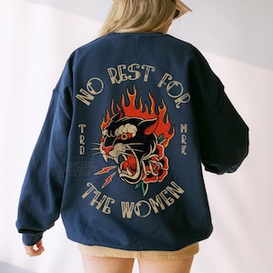 May include: Navy blue sweatshirt with a graphic of a black panther, red rose, and flames. The text "NO REST FOR THE WOMEN" is arched above the graphic. The sweatshirt is worn by a person wearing a tan baseball cap.