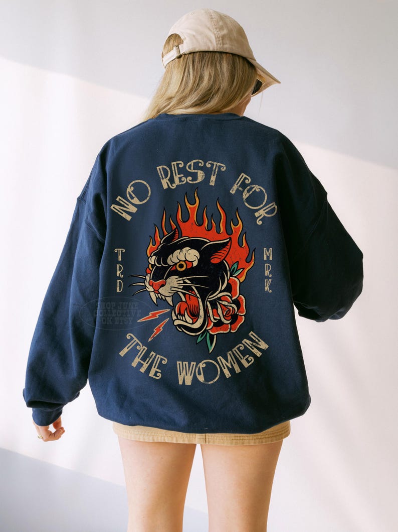 May include: Navy blue sweatshirt with a graphic of a black panther, red rose, and flames. The text "NO REST FOR THE WOMEN" is arched above the graphic. The sweatshirt is worn by a person wearing a tan baseball cap.