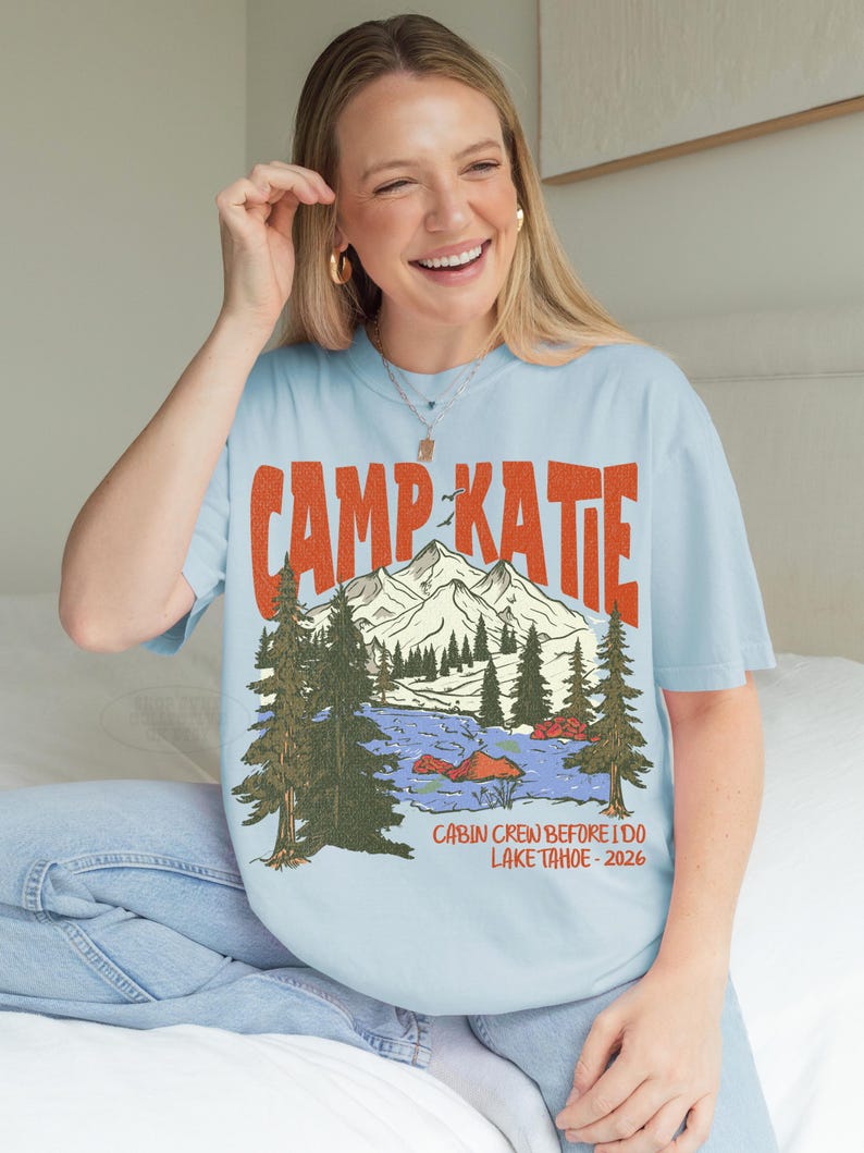 May include: Light blue t-shirt with the text "CAMP KATIE" in orange, above a mountain and lake graphic. The graphic includes trees and the text "CABIN CREW BEFORE I DO LAKE TAHOE - 2026". The shirt is worn by a person.