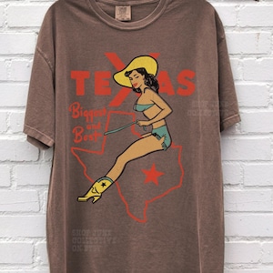 Op de afbeelding: Bruin t-shirt met een retro ontwerp van een vrouw met een cowboyhoed en laarzen, zittend op een Texas-omtrek. Het woord "TEXAS" is rood, met de tekst "Biggest and Best" eronder. Het t-shirt heeft een losse pasvorm.
