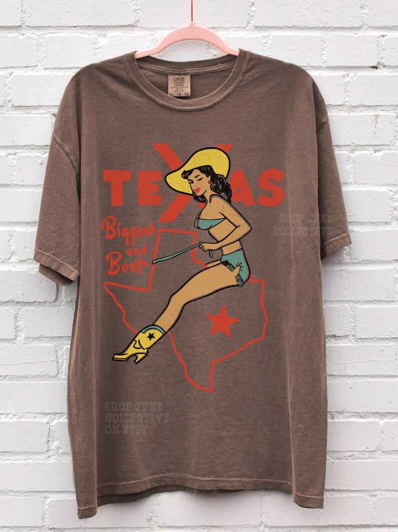 Op de afbeelding: Bruin t-shirt met een retro ontwerp van een vrouw met een cowboyhoed en laarzen, zittend op een Texas-omtrek. Het woord "TEXAS" is rood, met de tekst "Biggest and Best" eronder. Het t-shirt heeft een losse pasvorm.