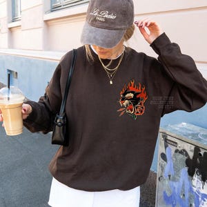 May include: A brown sweatshirt with a graphic of a black panther with red flames and a rose. The sweatshirt is worn with a gray baseball cap that says "La Po&eacute;sie PARIS". A small black purse is worn over the shoulder.