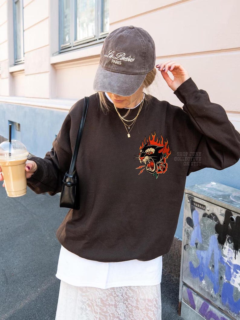 May include: A brown sweatshirt with a graphic of a black panther with red flames and a rose. The sweatshirt is worn with a gray baseball cap that says "La Po&eacute;sie PARIS". A small black purse is worn over the shoulder.