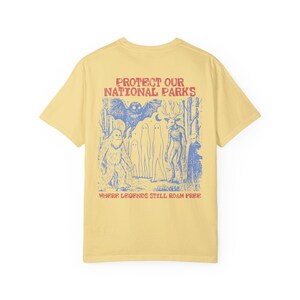 May include: A pale yellow short-sleeve t-shirt featuring a blue graphic print on the back. The design depicts a forest scene with a large owl, a sasquatch, ghosts, and a deer-like creature. Text reads: PROTECT OUR NATIONAL PARKS WHERE LEGENDS STILL ROAM FREE.