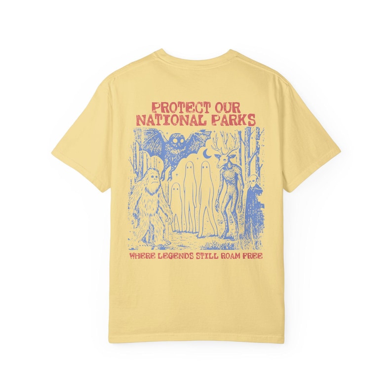 May include: A pale yellow short-sleeve t-shirt featuring a blue graphic print on the back. The design depicts a forest scene with a large owl, a sasquatch, ghosts, and a deer-like creature. Text reads: PROTECT OUR NATIONAL PARKS WHERE LEGENDS STILL ROAM FREE.