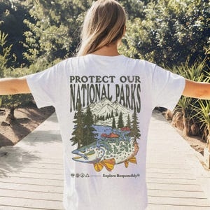 May include: White t-shirt with a graphic print of a mountain scene with a fish and the text "Protect Our National Parks" and "Explore Responsibly".