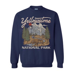 May include: A navy blue crewneck sweatshirt featuring a graphic print of Yellowstone National Park. The design includes a brown bison, a winding river, green pine trees, and grey mountains under a red sun, with the text Yellowstone National Park.