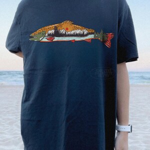 Puede incluir: Camiseta azul marino con un gr&aacute;fico detallado de un pez que contiene una escena de monta&ntilde;a y puesta de sol. El dise&ntilde;o del pez incorpora tonos de naranja, marr&oacute;n, verde y rojo. La camiseta es de corte holgado.