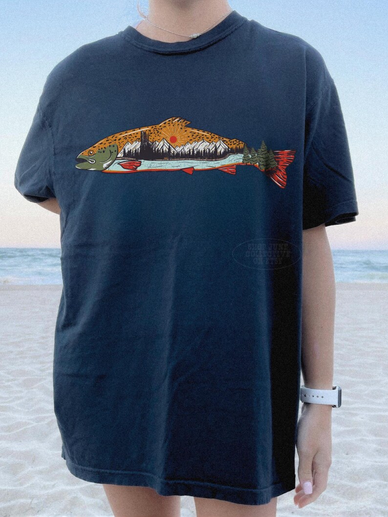 Puede incluir: Camiseta azul marino con un gr&aacute;fico detallado de un pez que contiene una escena de monta&ntilde;a y puesta de sol. El dise&ntilde;o del pez incorpora tonos de naranja, marr&oacute;n, verde y rojo. La camiseta es de corte holgado.